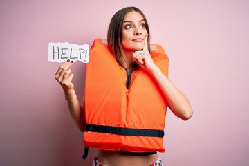 Young beautiful brunette woman wearing lifejacket holding paper with help word message serious face...