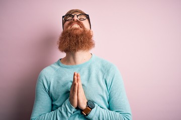 Handsome Irish redhead man with beard wearing glasses over pink isolated background begging and...