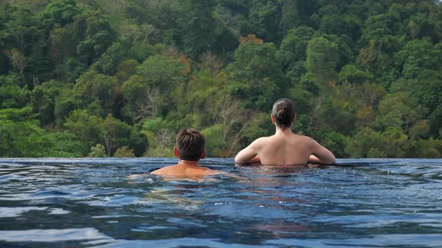 muscular guy swims to large pool edge kisses girlfriend and looks at pictorial hilly landscape with dense forests backside view