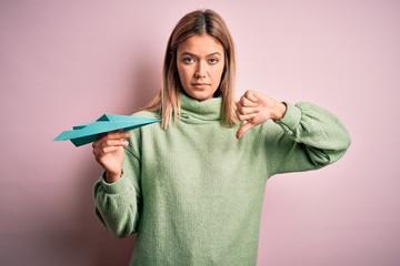Young beautiful woman holding paper airplane standing over isolated yellow background with angry face, negative sign showing dislike with thumbs down, rejection concept - obrazy, fototapety, plakaty