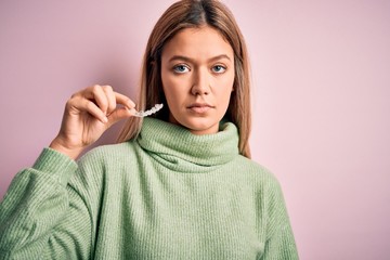 Young beautiful woman holding aligner standing over isolated pink background with a confident...