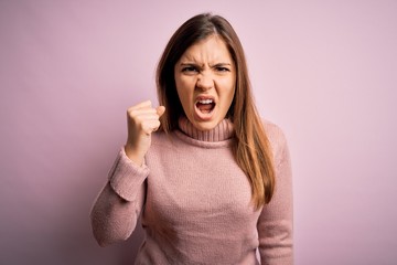 Beautiful young woman wearing turtleneck sweater over pink isolated background angry and mad raising fist frustrated and furious while shouting with anger. Rage and aggressive concept.