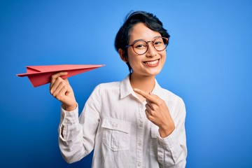 Young beautiful chinese woman holding paper airplane over isolated blue background very happy pointing with hand and finger