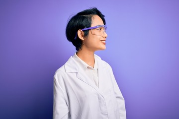 Young beautiful asian scientist girl wearing coat and glasses over purple background looking away to side with smile on face, natural expression. Laughing confident. - obrazy, fototapety, plakaty