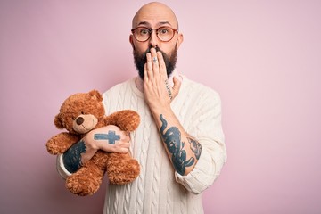 Handsome bald man with beard and tattoo holding teddy bear over isolated pink background cover...