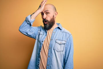 Handsome bald man with beard wearing casual denim jacket and striped t-shirt surprised with hand on...