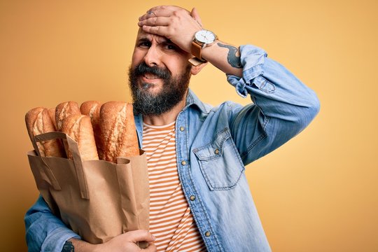 Handsome bald man with beard holding paper bag with bread over yellow background stressed with hand on head, shocked with shame and surprise face, angry and frustrated. Fear and upset for mistake.