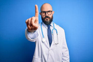 Handsome bald doctor man with beard wearing glasses and stethoscope over blue background Pointing with finger up and angry expression, showing no gesture