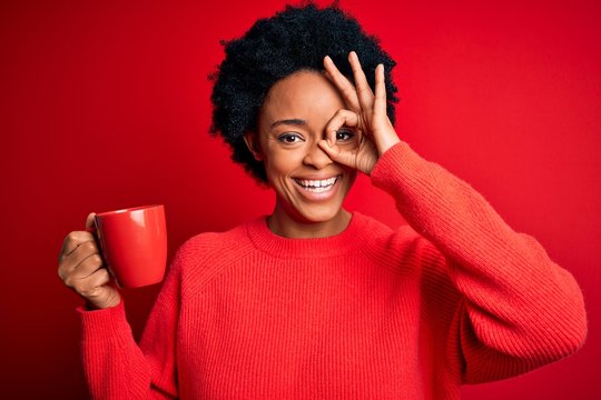 African American afro woman with curly hair drinking cup of coffee over red background with happy face smiling doing ok sign with hand on eye looking through fingers