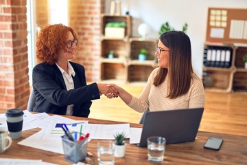 Two beautiful businesswomen smiling happy and confident. Sitting with smile on face working together using laptop shaking hands for agreement at the office