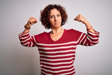 Middle age beautiful curly hair woman wearing casual striped sweater over white background showing arms muscles smiling proud. Fitness concept.