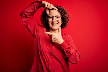 Middle age beautiful curly hair woman wearing casual shirt and glasses over red background smiling making frame with hands and fingers with happy face. Creativity and photography concept.