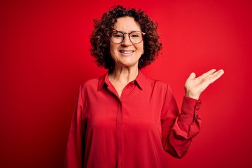 Middle age beautiful curly hair woman wearing casual shirt and glasses over red background smiling cheerful presenting and pointing with palm of hand looking at the camera.
