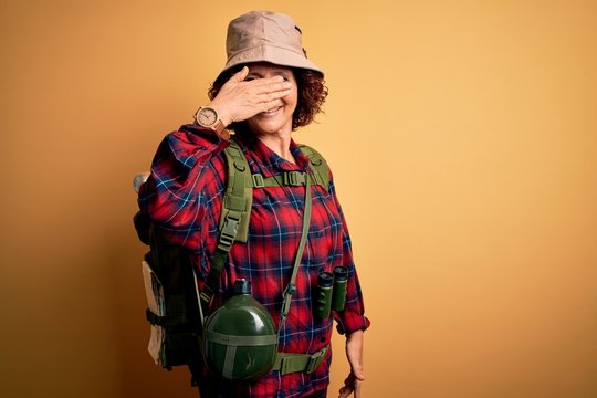 Middle age curly hair hiker woman hiking wearing backpack and water canteen using binoculars smiling and laughing with hand on face covering eyes for surprise. Blind concept.