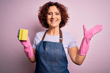 Middle age curly hair woman cleaning doing housework wearing apron and gloves using scourer very happy and excited, winner expression celebrating victory screaming with big smile and raised hands