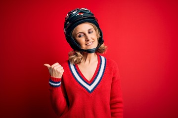 Middle age beautiful blonde motorcyclist woman wearing moto helmet over red background smiling with happy face looking and pointing to the side with thumb up.