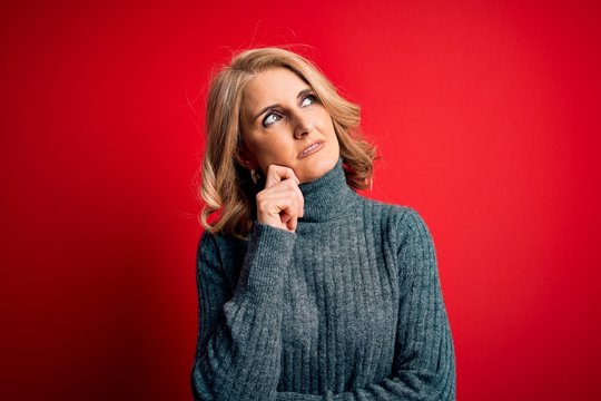 Middle age beautiful blonde woman wearing casual turtleneck sweater over red background with hand on chin thinking about question, pensive expression. Smiling with thoughtful face. Doubt concept.