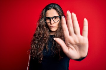 Young beautiful woman with curly hair wearing sweater and glasses over red background doing stop sing with palm of the hand. Warning expression with negative and serious gesture on the face.