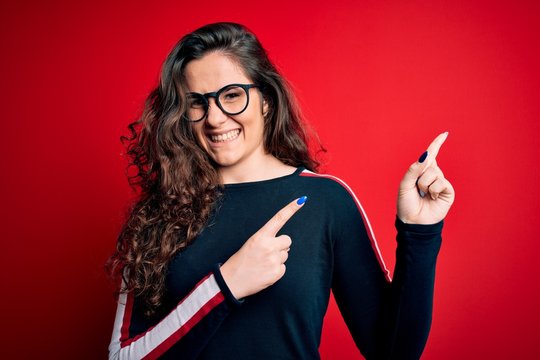 Young beautiful woman with curly hair wearing sweater and glasses over red background smiling and looking at the camera pointing with two hands and fingers to the side.