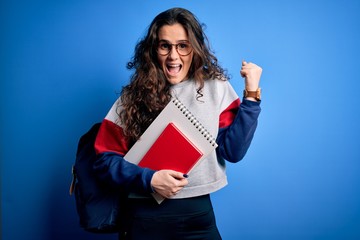 Young beautiful student woman with curly hair wearing backpack holding book and notebook screaming proud and celebrating victory and success very excited, cheering emotion