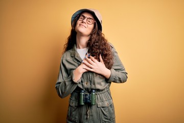 Young beautiful tourist woman on vacation wearing explorer hat and binoculars smiling with hands on chest with closed eyes and grateful gesture on face. Health concept.