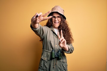 Young beautiful tourist woman on vacation wearing explorer hat and binoculars smiling looking to the camera showing fingers doing victory sign. Number two.