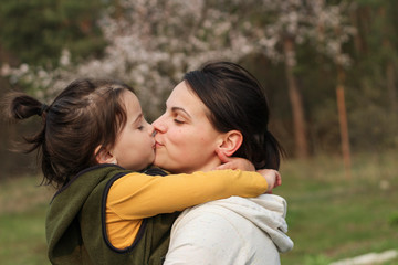 Beautiful mother and daughter hugging and kissing in casual clothes. Warm baby hug on the background of a blurry flowering tree
