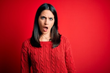 Fototapeta premium Young brunette woman with blue eyes wearing casual sweater over isolated red background In shock face, looking skeptical and sarcastic, surprised with open mouth