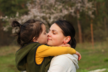 Fototapeta premium Beautiful mother and daughter hugging and kissing in casual clothes. Warm baby hug on the background of a blurry flowering tree
