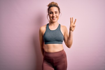 Young beautiful blonde sportswoman doing sport wearing sportswear over pink background showing and pointing up with fingers number three while smiling confident and happy.