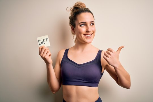 Young beautiful blonde sportswoman wearing sportswear holding reminder with diet message pointing and showing with thumb up to the side with happy face smiling