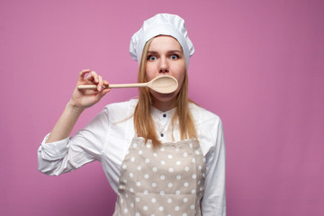 beautiful surprised girl cook in kitchen clothes with a spoon closes her mouth on a pink background, woman housewife with kitchen items