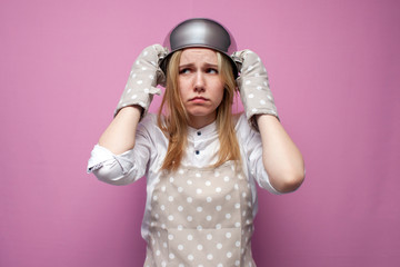 unhappy girl cook in kitchen clothes with a pan on her head is sad on a colored background, woman is a housewife with kitchen items in stress