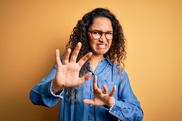 Beautiful woman with curly hair wearing casual denim shirt and glasses over yellow background...