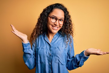 Beautiful woman with curly hair wearing casual denim shirt and glasses over yellow background...