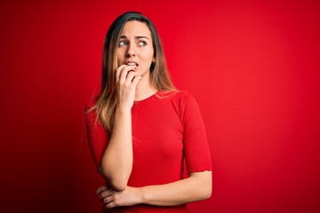 Young beautiful blonde woman with blue eyes wearing casual t-shirt over red background looking stressed and nervous with hands on mouth biting nails. Anxiety problem.