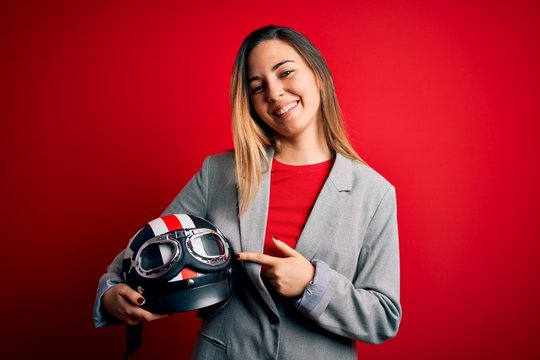 Young beautiful blonde motorcyclist woman holding motorcycle helmet over red background very happy pointing with hand and finger