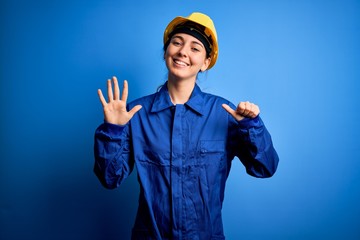 Young beautiful worker woman with blue eyes wearing security helmet and uniform showing and pointing up with fingers number six while smiling confident and happy.