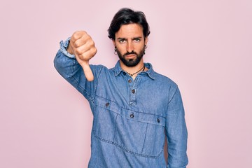 Young handsome hispanic bohemian man wearing hippie style over pink background looking unhappy and angry showing rejection and negative with thumbs down gesture. Bad expression.