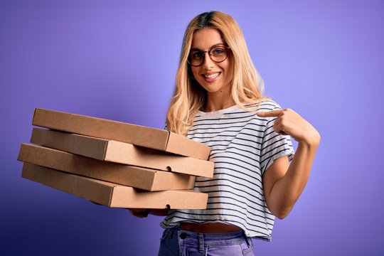 Young beautiful blonde woman holding boxes of italian pizza over isolated purple background with surprise face pointing finger to himself