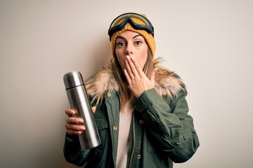Young beautiful brunette skier woman wearing ski goggles drinking thermo with coffe cover mouth...