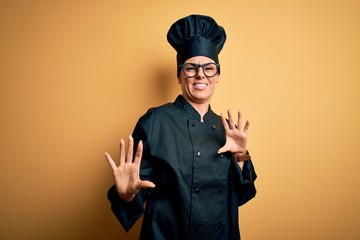 Young beautiful brunette chef woman wearing cooker uniform and hat over yellow background disgusted expression, displeased and fearful doing disgust face because aversion reaction. With hands raised