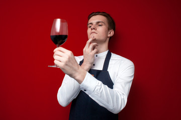 professional thoughtful sommelier holds a glass of red wine and thinks against a red background,...