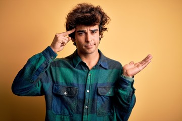 Young handsome man wearing casual shirt standing over isolated yellow background confused and annoyed with open palm showing copy space and pointing finger to forehead. Think about it.