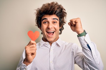 Young handsome man holding paper heart standing over isolated white background screaming proud and celebrating victory and success very excited, cheering emotion