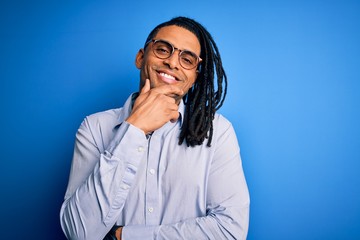 Young handsome african american man with dreadlocks wearing casual shirt and glasses looking confident at the camera with smile with crossed arms and hand raised on chin. Thinking positive.
