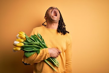 Young african american afro romantic man with dreadlocks holding bouquet of yellow tulips angry and mad screaming frustrated and furious, shouting with anger. Rage and aggressive concept.