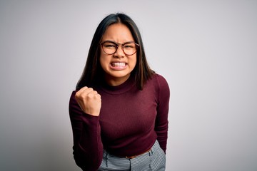 Beautiful asian business woman wearing casual sweater and glasses over white background angry and mad raising fist frustrated and furious while shouting with anger. Rage and aggressive concept.