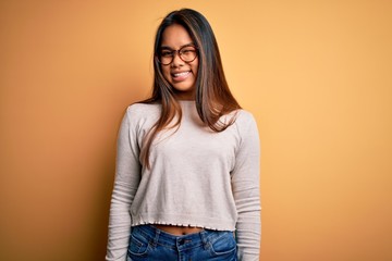 Young beautiful asian girl wearing casual sweater and glasses over yellow background winking...