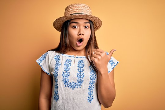 Young beautiful asian girl wearing casual t-shirt and hat standing over yellow background Surprised pointing with hand finger to the side, open mouth amazed expression.